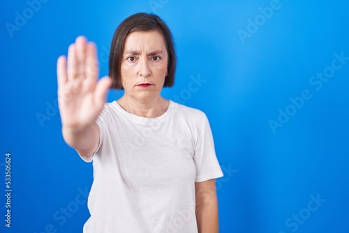 Middle age hispanic woman standing over blue background doing stop sing with palm of the hand. warning expression with negative and serious gesture on the face. © Krakenimages.com