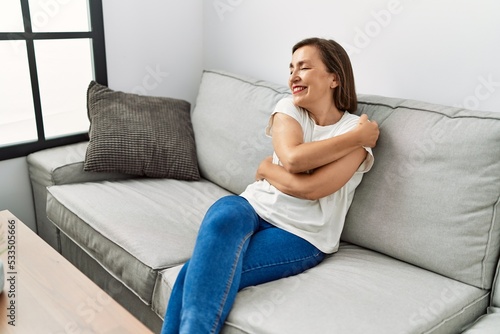 Middle age hispanic woman smiling sitting on the sofa hugging herself at home