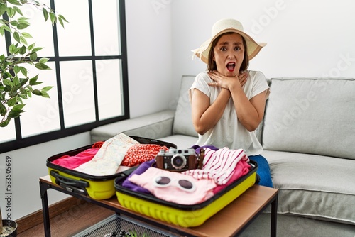 Beautiful middle age hispanic woman packing summer clothes in suitcase shouting and suffocate because painful strangle. health problem. asphyxiate and suicide concept.