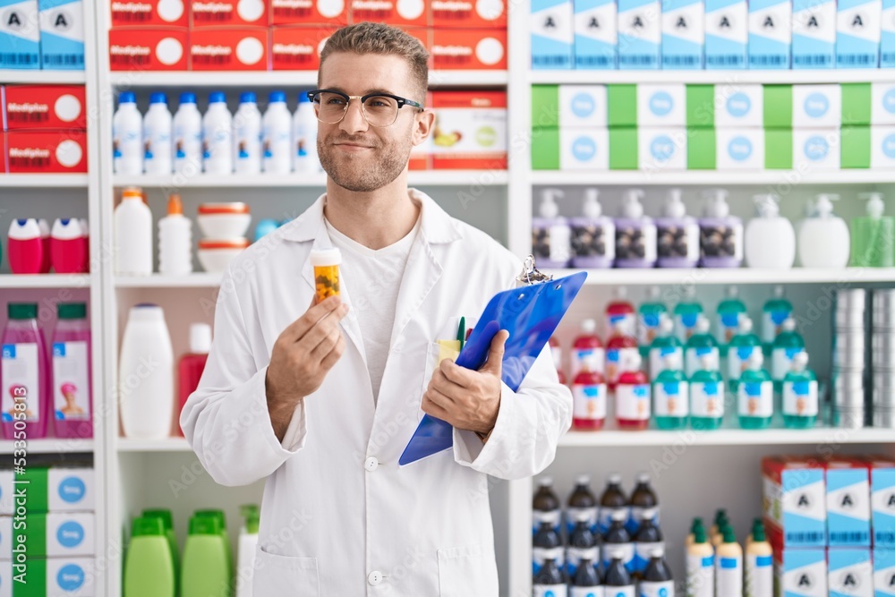 Young caucasian man working at pharmacy drugstore holding pills smiling looking to the side and staring away thinking.