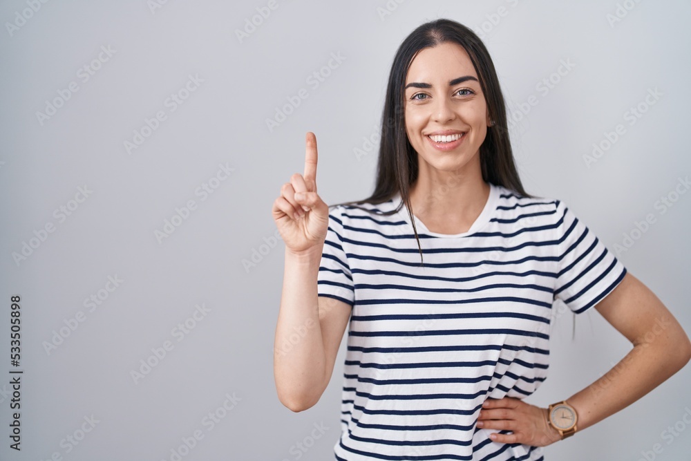 Young brunette woman wearing striped t shirt showing and pointing up with finger number one while smiling confident and happy. Young brunette woman wearing striped t shirt showing and pointing up with finger number one while smiling confident and happy.