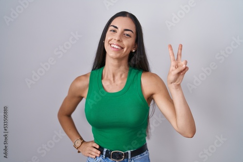 Young woman standing over isolated background smiling looking to the camera showing fingers doing victory sign. number two.