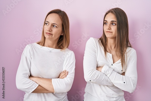 Middle age mother and young daughter standing over pink background looking to the side with arms crossed convinced and confident