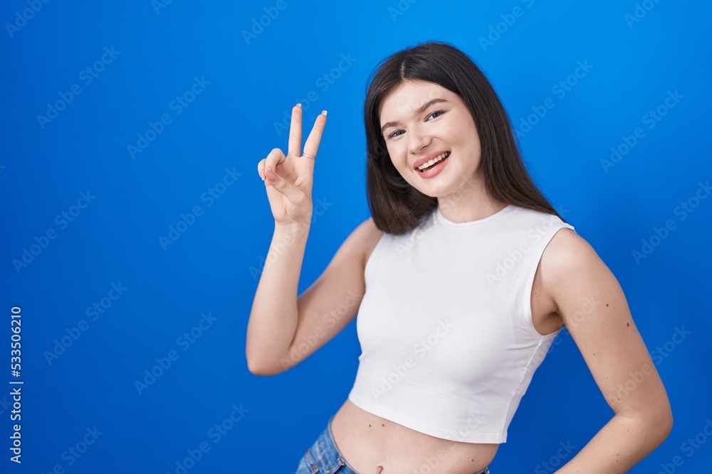 Young caucasian woman standing over blue background smiling looking to the camera showing fingers doing victory sign. number two. Young caucasian woman standing over blue background smiling looking to the camera showing fingers doing victory sign. number two.