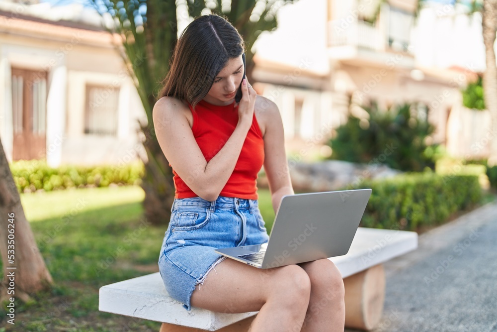 Young caucasian woman using laptop talking on smartphone sitting on bench at park Young caucasian woman using laptop talking on smartphone sitting on bench at park