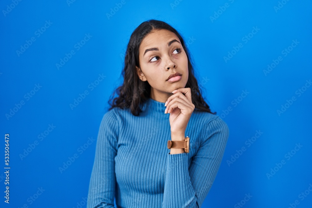 Young brazilian woman standing over blue isolated background with hand on chin thinking about question, pensive expression. smiling with thoughtful face. doubt concept. Young brazilian woman standing over blue isolated background with hand on chin thinking about question, pensive expression. smiling with thoughtful face. doubt concept.