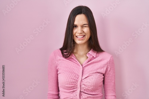Young hispanic woman standing over pink background winking looking at the camera with sexy expression, cheerful and happy face.