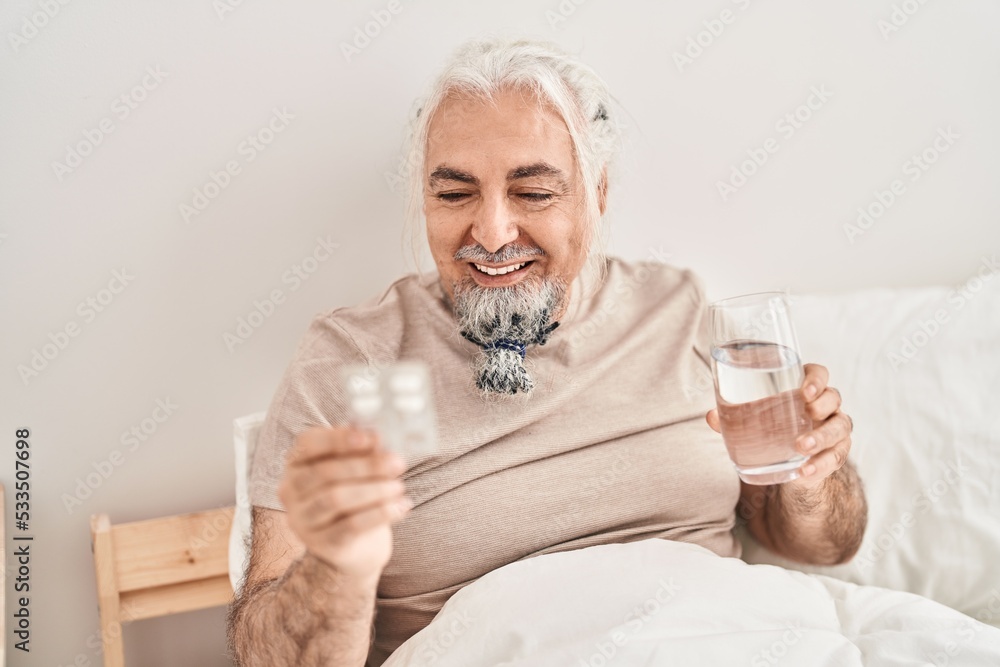 Middle age grey-haired man holding pills tablet and glass of water sitting on bed at bedroom Middle age grey-haired man holding pills tablet and glass of water sitting on bed at bedroom