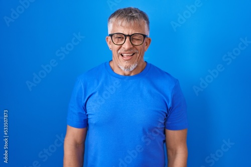 Hispanic man with grey hair standing over blue background winking looking at the camera with sexy expression, cheerful and happy face.