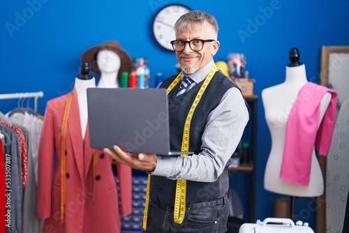 Middle age grey-haired man tailor smiling confident using laptop at clothing factory
