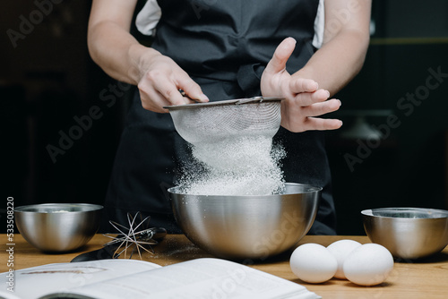 Foto Cropped image of female pastry chef sifting flour for homemade baking, woman in apron holding sieve surrounded by baking ingredients
