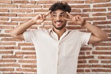 Arab man with beard standing over bricks wall background doing peace symbol with fingers over face, smiling cheerful showing victory