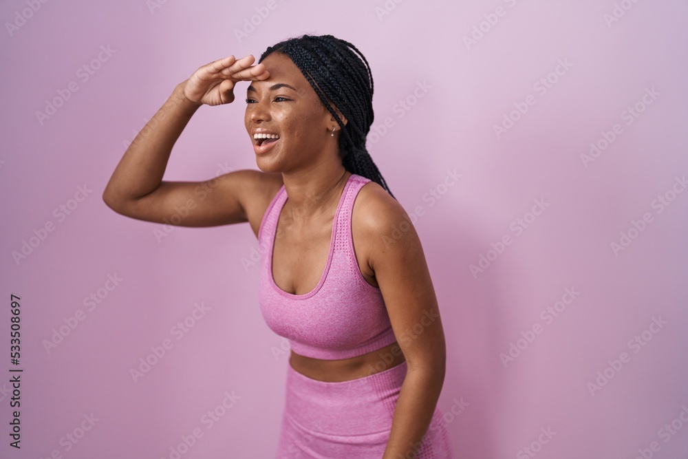 African american woman with braids wearing sportswear over pink background very happy and smiling looking far away with hand over head. searching concept. African american woman with braids wearing sportswear over pink background very happy and smiling looking far away with hand over head. searching concept.