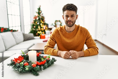 Arab young man sitting on the table by christmas tree relaxed with serious expression on face. simple and natural looking at the camera.