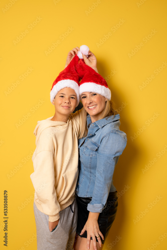 Fototapeta premium Pretty young mom and her handsome child son together wearing Santa hats. Isolated on yellow.