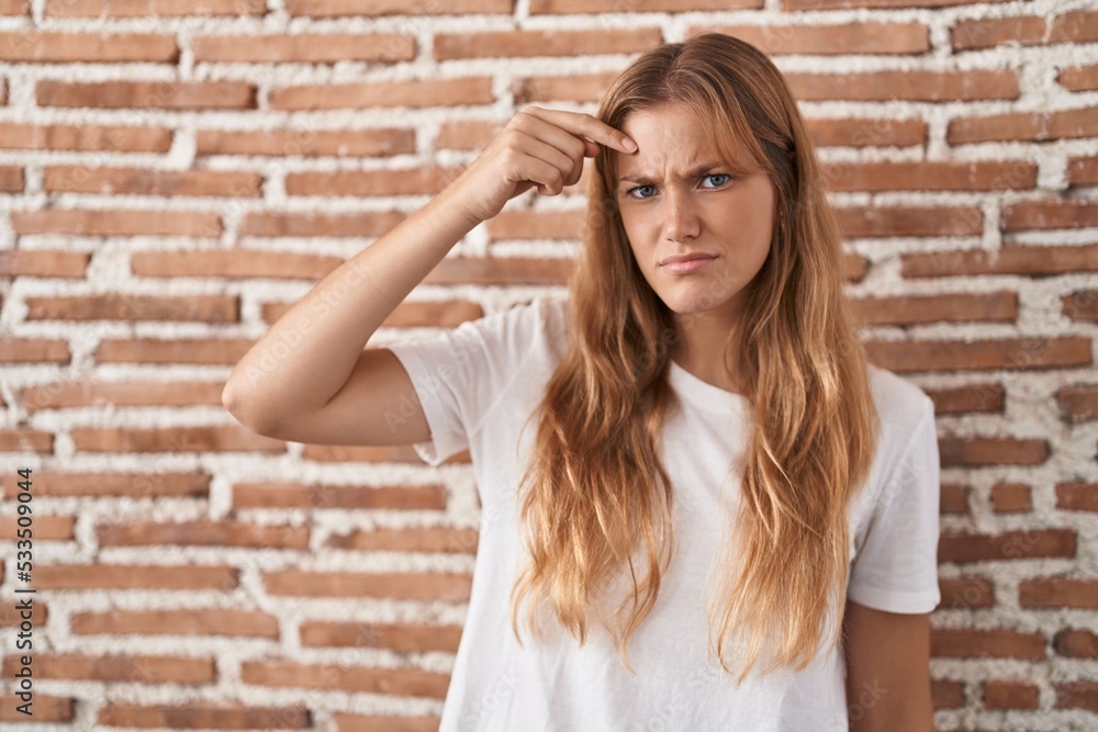 Young caucasian woman standing over bricks wall pointing unhappy to pimple on forehead, ugly infection of blackhead. acne and skin problem Young caucasian woman standing over bricks wall pointing unhappy to pimple on forehead, ugly infection of blackhead. acne and skin problem