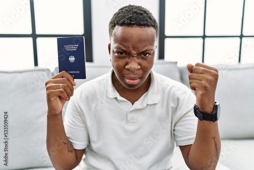 Young african man holding deutschland passport annoyed and frustrated shouting with anger, yelling crazy with anger and hand raised