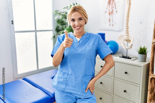 Beautiful blonde physiotherapist woman working at pain recovery clinic doing happy thumbs up gesture with hand. approving expression looking at the camera showing success.