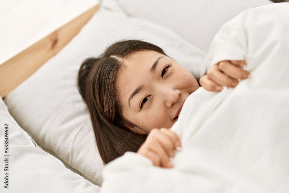 Young chinese girl covering with bedsheet lying on the bed at bedroom.