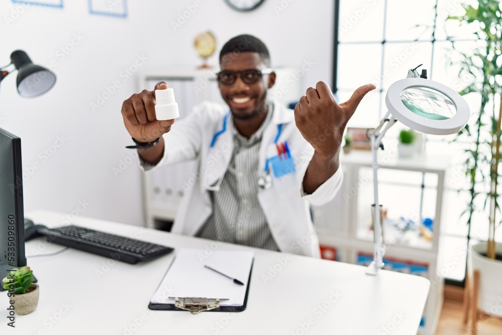 Young african american doctor man wearing doctor uniform holding prescription pills at the clinic pointing thumb up to the side smiling happy with open mouth