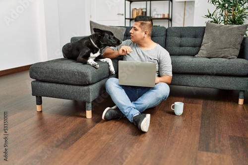 Young hispanic man using laptop sitting on the floor with dog at home.