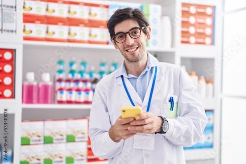 Young hispanic man pharmacist using smartphone working at pharmacy