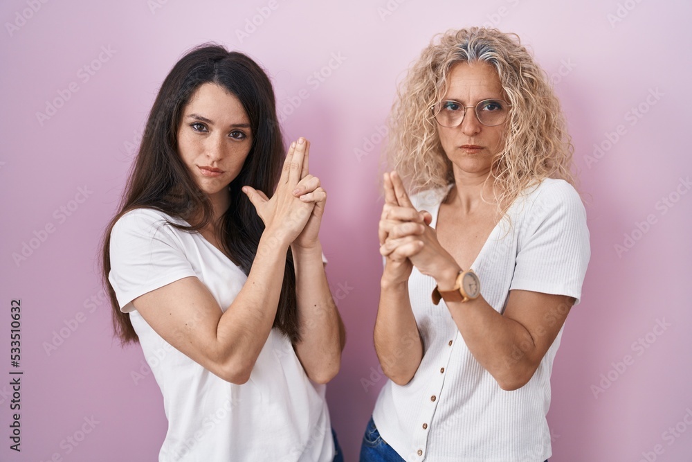 Mother and daughter standing together over pink background holding symbolic gun with hand gesture, playing killing shooting weapons, angry face Mother and daughter standing together over pink background holding symbolic gun with hand gesture, playing killing shooting weapons, angry face