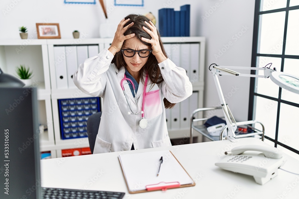 Young doctor woman wearing doctor uniform and stethoscope at the clinic suffering from headache desperate and stressed because pain and migraine. hands on head. Young doctor woman wearing doctor uniform and stethoscope at the clinic suffering from headache desperate and stressed because pain and migraine. hands on head.