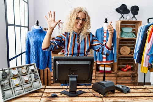 Middle age blonde woman working as manager at retail boutique showing and pointing up with fingers number six while smiling confident and happy.
