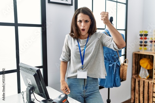 Young brunette woman working as manager at retail boutique angry and mad raising fist frustrated and furious while shouting with anger. rage and aggressive concept.