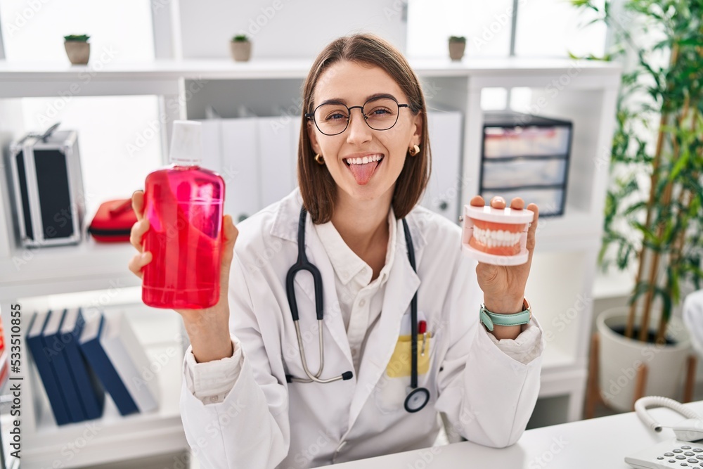 Young caucasian dentist woman holding denture and mouthwash sticking tongue out happy with funny expression. Young caucasian dentist woman holding denture and mouthwash sticking tongue out happy with funny expression.