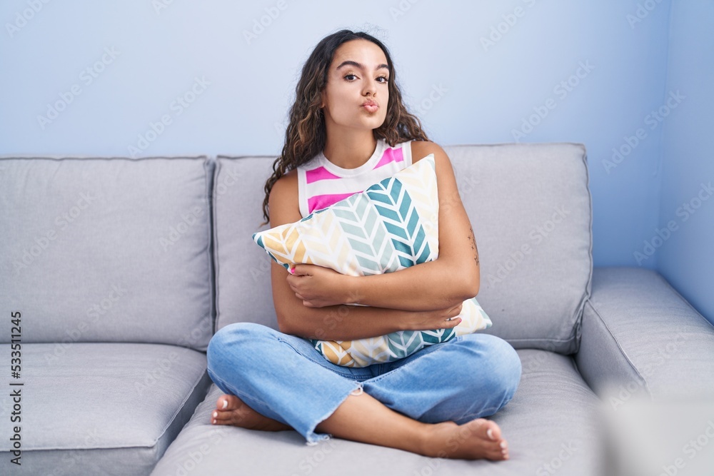 Young hispanic woman sitting on the sofa at home looking at the camera blowing a kiss being lovely and sexy. love expression.