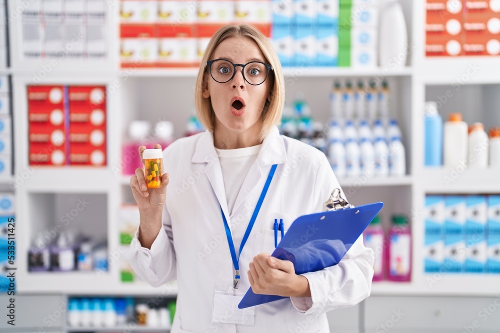 Young caucasian woman working at pharmacy drugstore holding pills afraid and shocked with surprise and amazed expression, fear and excited face.