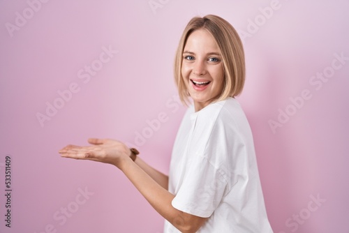 Young caucasian woman standing over pink background pointing aside with hands open palms showing copy space, presenting advertisement smiling excited happy