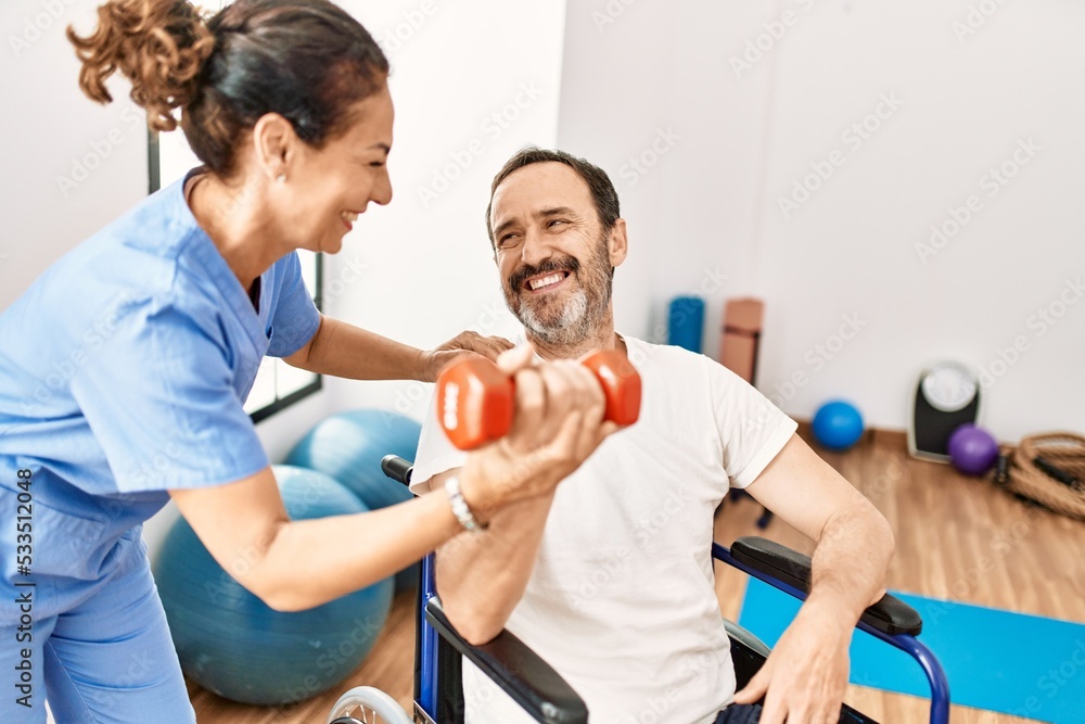 Obraz premium Middle age man and woman having rehab session using dumbbell sitting on wheelchair at physiotherapy clinic