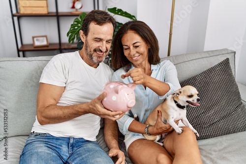 Middle age man and woman couple holding piggy bank sitting on sofa with dog at home