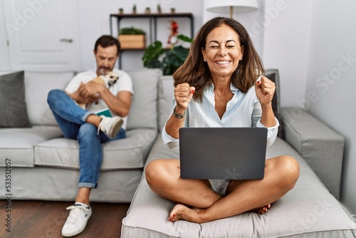 Hispanic middle age couple at home, woman using laptop excited for success with arms raised and eyes closed celebrating victory smiling. winner concept.