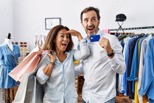 Hispanic middle age couple holding shopping bags and credit card very happy and smiling looking far away with hand over head. searching concept.