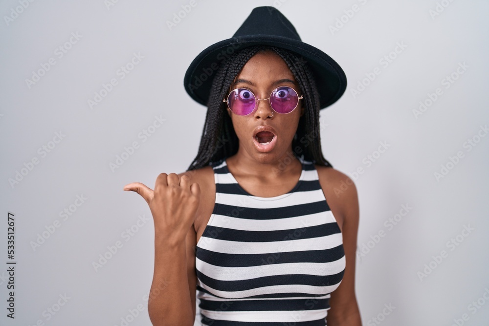 Young african american with braids wearing hat and sunglasses surprised pointing with hand finger to the side, open mouth amazed expression. Young african american with braids wearing hat and sunglasses surprised pointing with hand finger to the side, open mouth amazed expression.