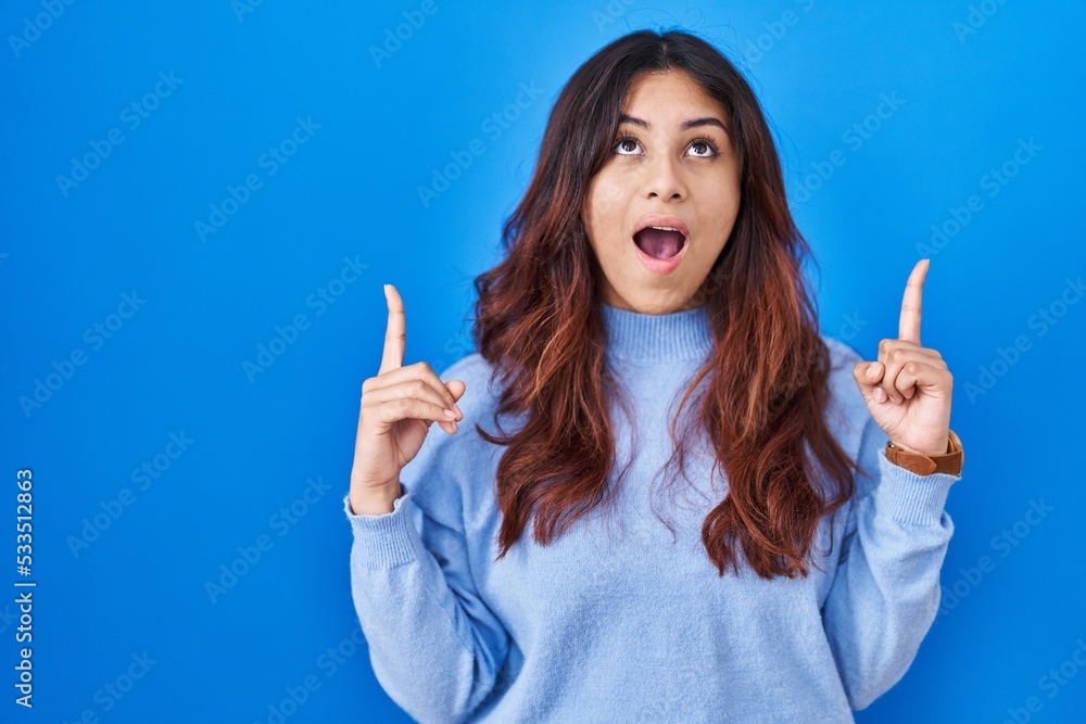 Hispanic young woman standing over blue background amazed and surprised looking up and pointing with fingers and raised arms. Hispanic young woman standing over blue background amazed and surprised looking up and pointing with fingers and raised arms.