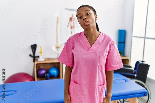 Young african american woman working at pain recovery clinic making fish face with lips  crazy and comical gesture. funny expression.
