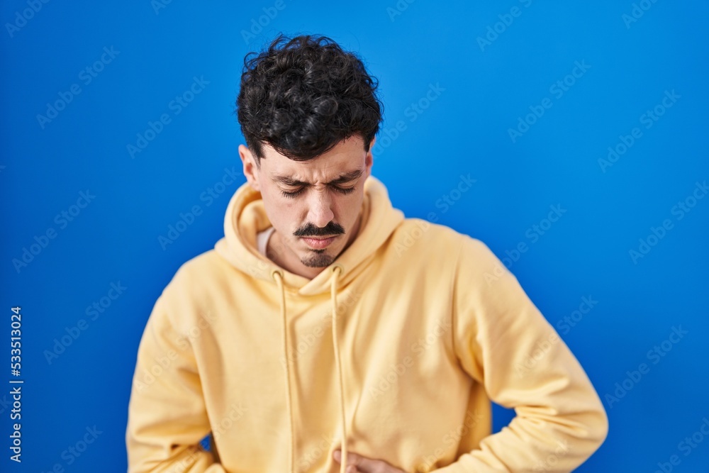 Fototapeta premium Hispanic man standing over blue background with hand on stomach because indigestion, painful illness feeling unwell. ache concept.