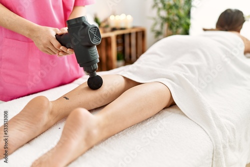 Young hispanic woman having legs massage with percussion gun at beauty center.