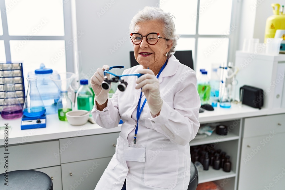Senior woman with grey hair working at scientist laboratory using magnifying glasses winking looking at the camera with sexy expression, cheerful and happy face. Senior woman with grey hair working at scientist laboratory using magnifying glasses winking looking at the camera with sexy expression, cheerful and happy face.