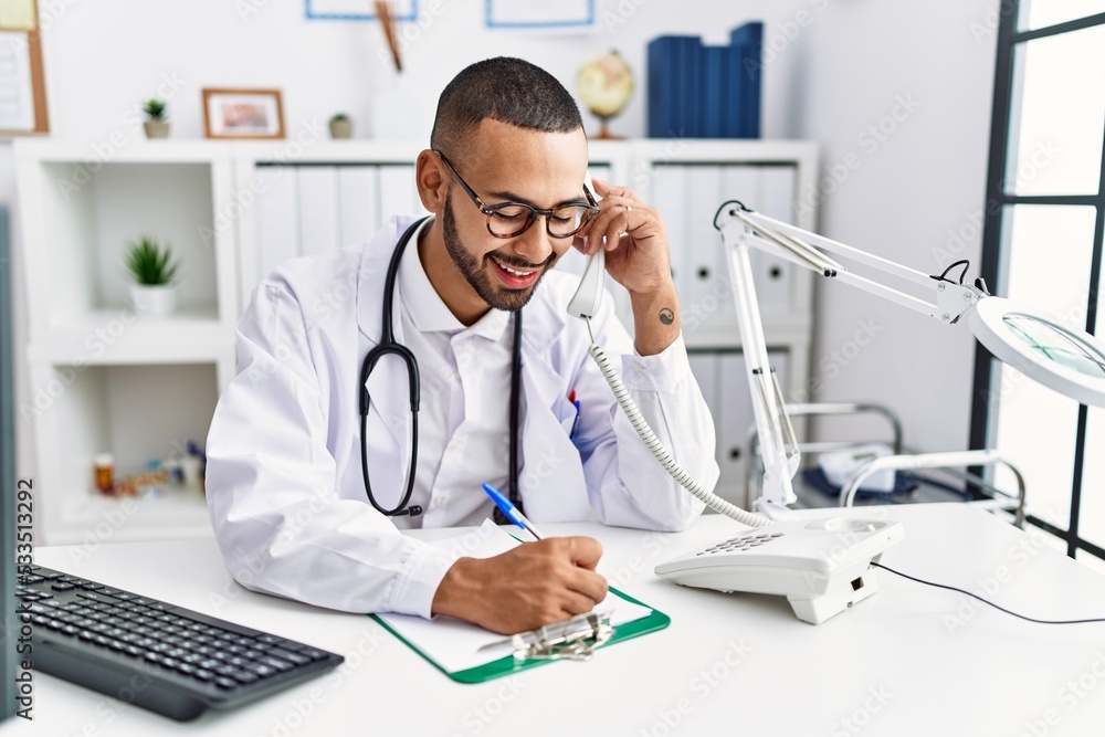 Young hispanic man wearing doctor uniform speaking on the phone at clinic