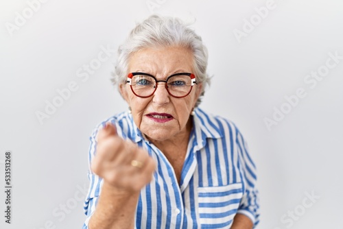 Senior woman with grey hair standing over white background angry and mad raising fist frustrated and furious while shouting with anger. rage and aggressive concept.