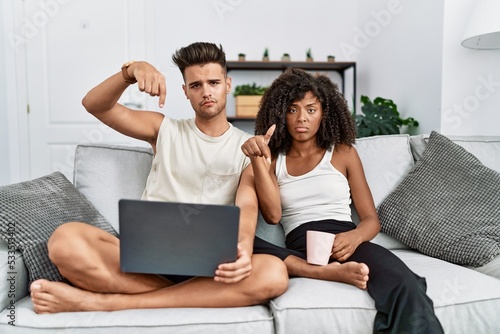 Young interracial couple using laptop at home sitting on the sofa pointing down looking sad and upset, indicating direction with fingers, unhappy and depressed.
