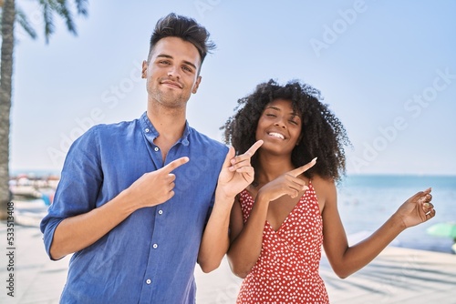 Young interracial couple outdoors on a sunny day smiling and looking at the camera pointing with two hands and fingers to the side.
