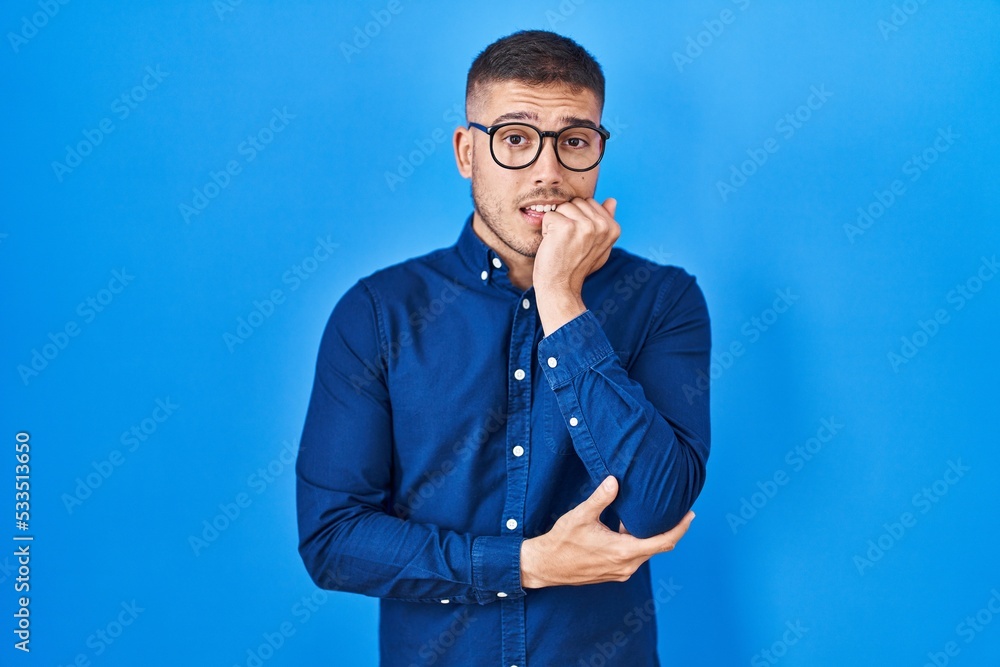 Young hispanic man wearing glasses over blue background looking stressed and nervous with hands on mouth biting nails. anxiety problem. Young hispanic man wearing glasses over blue background looking stressed and nervous with hands on mouth biting nails. anxiety problem.