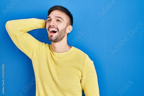 Hispanic man standing over blue background smiling confident touching hair with hand up gesture, posing attractive and fashionable